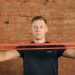 Adult male using a resistance band for fitness training against a brick wall backdrop.